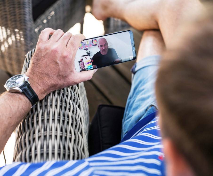 een man zit buiten op de stoel in zomerkleding en is via zijn telefoon met een coach aan het videobellen