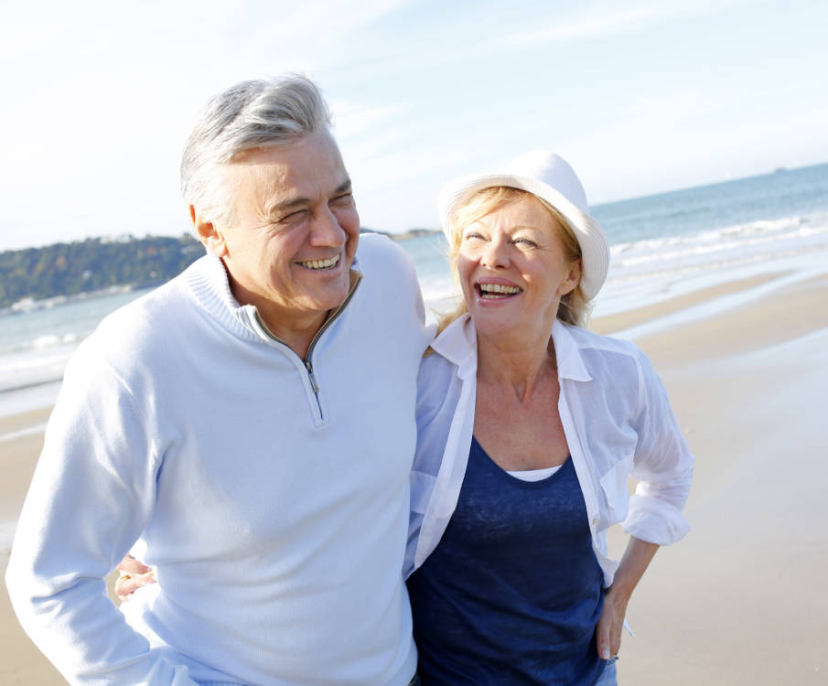Man en vrouw lopen arm in arm op een zonnig strand. Ze lachen.