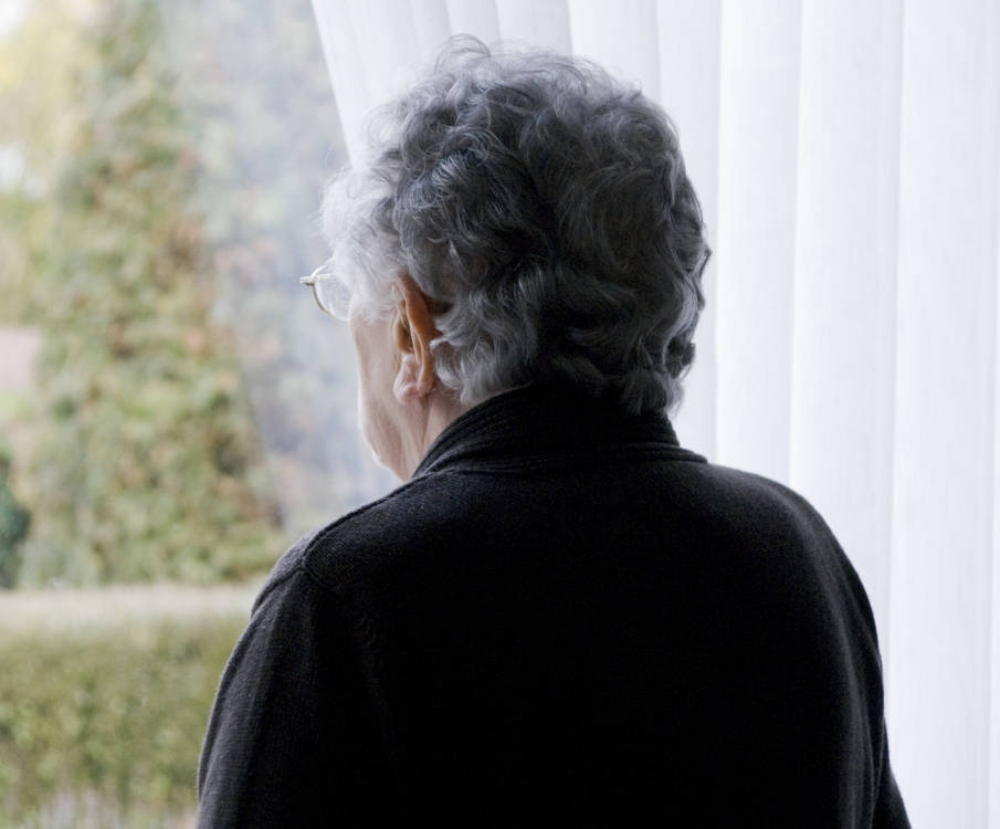 Een oude dame met grijs haar kijkt uit het raam naar buiten. We zien haar rug.