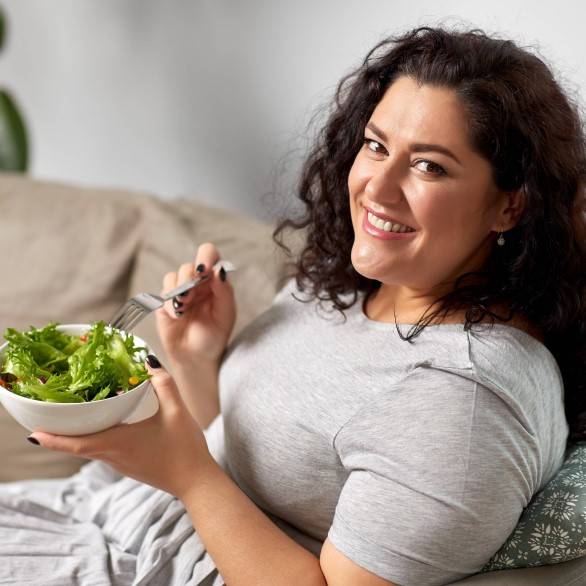 een vrouw met bruine krullenzit op de bank met een bak gezonde salade die ze aan het eten is