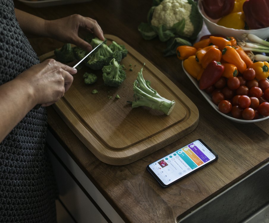 een vrouw snijdt broccoli op een houten plank voor de schaal met verse groenten en kijkt op de health coach app