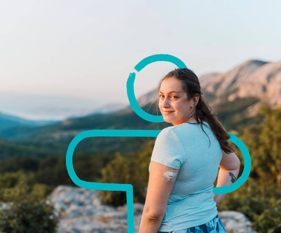 Een vrouw staat op een berg met veel bomen. Ze heeft een blauw shirt aan en glucosesensor op haar arm. Ze draait zich om richting de camera en lacht.