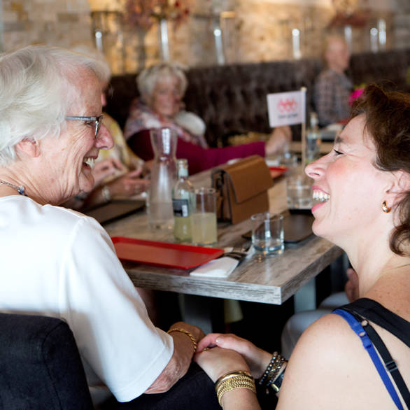 Oudere vrouw zit aan tafel met andere alleenstaande ouderen en maakt praatje met begeleider