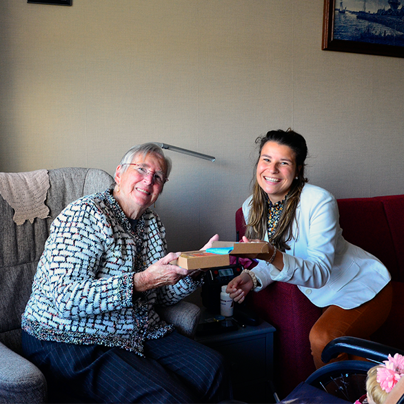 Oudere in stoel en medewerker Mediq lachen met koek gegeven door Stichting met je hart. 