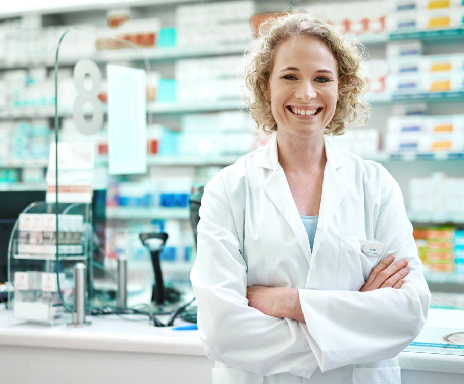 Vrouwelijke apotheker met een witte jas aan lacht en staat voor de apothekersbalie