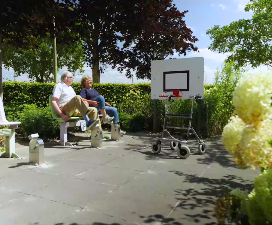 Twee ouderen zitten op een bankje op een plein. Er is een lage basketbalring waar een rollator voor staat.